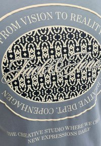 Μπλε ύφασμα με κεντρικό οβάλ σχέδιο που περιλαμβάνει μπεζ γεωμετρικά σχήματα, γραφικό κείμενο "Jack & Jones" σε πλάγια γραφή και περιμετρικά συνθήματα για τη δημιουργικότητα και την Κοπεγχάγη.