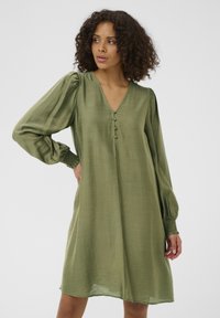 Robe mi-longue couleur vert olive avec un décolleté en V, dotée d'un devant boutonné et de manches longues bouffantes avec des poignets smockés. Texture lisse.