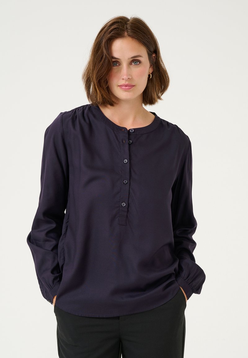 Blouse bleu marine à boutons avec un col rond, des manches longues et des détails froncés aux épaules. Le tissu semble lisse et léger.