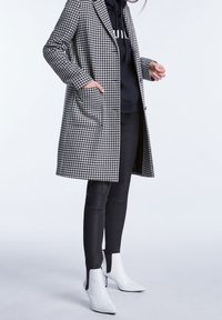 Mujer vestida con un abrigo de pata de gallo en blanco y negro, sudadera negra, leggings negros y botines de tacón blanco, de pie contra un fondo liso.