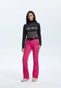 Black zip-up top with gold geometric pattern; bright pink pants with belt and side pockets; model standing on plain background.