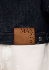 Σκούρο μπλε τζιν μπουφάν με καφέ δερμάτινο ταμπελάκι με την ένδειξη "MAX & Co." και ορατό μεταλλικό κουμπί σε φόντο λευκού παντελονιού.