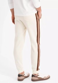 Pantalones de chándal cremosos con franjas laterales marrones, ajuste ceñido, puños acanalados y una textura suave. Se llevan con zapatillas blancas y marrones.