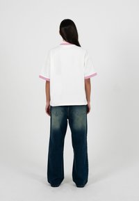 Person mit langen dunklen Haaren steht mit dem Rücken zur Kamera, trägt ein weißes Kurzarmhemd mit pinkfarbenem Rand und lockere dunkelblaue Jeans.