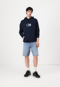 Felpa blu navy con un design grafico sul petto, abbinata a pantaloni shorts in denim chiaro e sneakers nere. Calzini bianchi in vista.