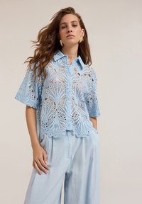 Ljusblå kortärmad skjorta i intrikat spets, med ett blommönster, knappstängning och vågig fåll. Matchas med ljusa byxor.