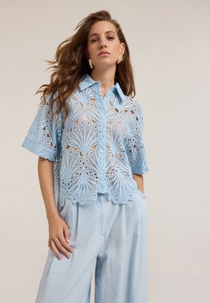 Camicia azzurra a maniche corte realizzata in pizzo intricato, con un motivo floreale, chiusura a bottoni e orlo scalloppato. Abbinata a pantaloni chiari coordinati.