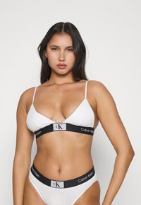 Calvin Klein bijela triangularna bralette s tankim trakama i crnim logom; usklađeni bijeli gaćice sličnog crnog loga.
