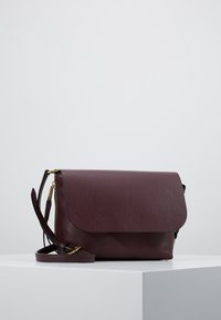Burgundská kožená crossbody taška s klopou, zlatým kováním a nastavitelným popruhem, ležící na světlém povrchu.