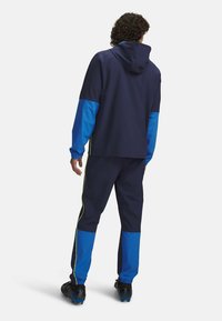 Navy tracksuit with blue accents, featuring a hooded jacket and tapered pants with neon yellow side stripes. Made of lightweight material.
