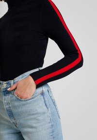 Marineblauwe coltrui met ribbelstructuur en rode strepen langs de mouwen, gecombineerd met lichtblauwe high-waisted spijkerbroek.
