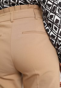 Beige skræddersyede bukser med baglommer, båret sammen med en langærmet bluse i sort og hvid med geometrisk mønster.