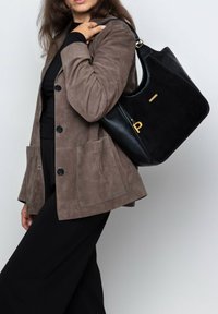 Sac à main en cuir noir avec des accents en suède, matériel doré et forme arrondie, porté avec une veste en suède marron dotée de grandes poches.