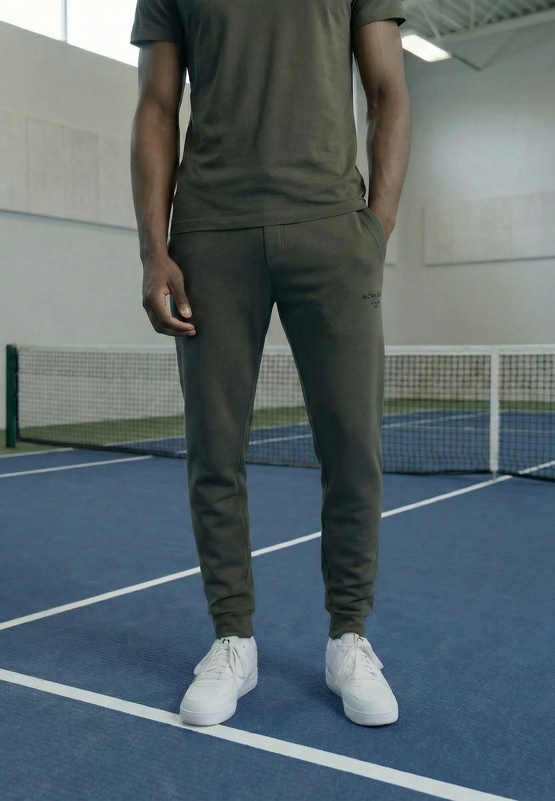 Mies, jolla on päällään tummanvihreä t-paita ja verkkarit valkoisilla lenkkareilla, seisoo sisätiloissa sinisellä tenniskentällä lähellä verkkoa.