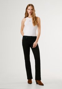 Jeans neri abbinati a un top senza maniche bianco, con un collo alto e un logo sottile, completati da stivali alla caviglia marroni con tacco.
