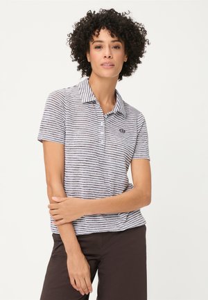 Donna con i capelli ricci che indossa una polo a maniche corte a righe grigio e bianco e pantaloni scuri, in piedi davanti a uno sfondo chiaro e semplice.