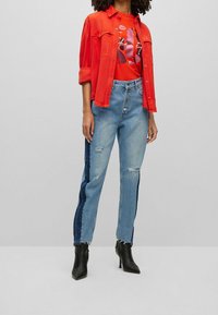 Czerwona koszula z denimu nałożona na czerwony t-shirt z nadrukiem, w połączeniu z jasnoniebieskimi dżinsami z ciemnym denimu bocznymi panelami i przetarciami. Czarne botki za kostkę.