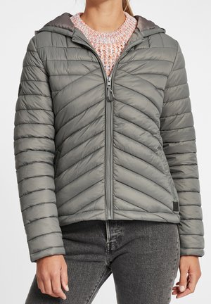 Femme portant une veste matelassée grise à fermeture éclair avec capuche, par-dessus un pull en tricot blanc et orange, associée à un jean gris foncé.
