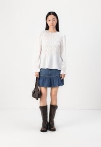 Hvit smock-bluse, dongerikjole med flere lag, brune støvletter til kneet og en liten mørk håndveske. Enkel design med volanger.