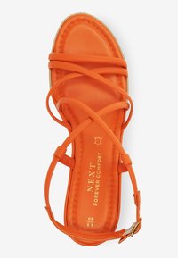 Sandale orange avec brides croisées, tige en cuir lisse, semelle plate et lettrage doré sur la semelle intérieure. Détail boucle réglable à la cheville.