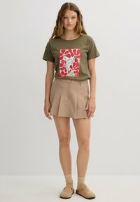 Camiseta verde oliva con un colorido estampado floral, combinada con una falda plisada beige. Cuenta con mangas cortas y un corte relajado.