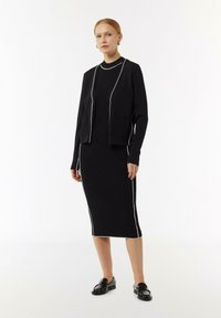 Schwarzes Strickkleid mit weißen Paspelierungen, kombiniert mit einer passenden schwarzen, kurzen Strickjacke. Das Model trägt schwarze Mokassins und steht vor einem einfarbigen Hintergrund.