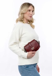 Borsa a tracolla rossa in pelle con patta curva, hardware dorato e tracolla regolabile, appoggiata contro un maglione bianco e soffice e jeans blu.