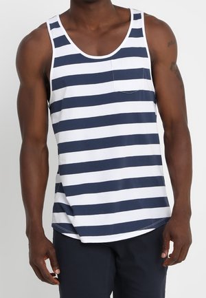 Mann trägt dunkelblau-weiß gestreiftes ärmelloses Tanktop mit kleiner Brusttasche, kombiniert mit dunkler Hose, steht vor schlichtem Hintergrund.