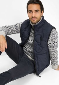 Marineblau gesteppte Weste über einem hellgrauen Pullover mit geometrischem Muster. Dunkle Hose rundet den Look ab, mit einer glatten Stofftextur.