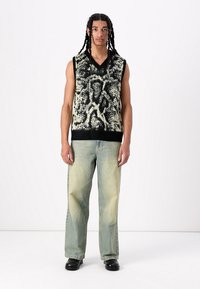 Wolblend, mouwloos, v-hals trui met een zwart en crèmekleurig abstract patroon. Draag het met lichtblauwe wijde jeans en zwarte schoenen.