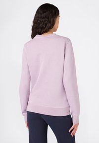 Felpa lilla con collo rotondo, maniche e orlo a coste. Tessuto morbido, vestibilità ampia e design leggermente più lungo sul retro. Abbinata a leggins scuri.