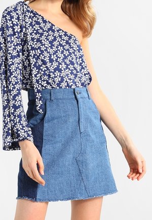 Frau trägt ein blaues florales Ein-Schulter-Top und einen hoch taillierten Jeansrock mit ausgefranstem Saum.