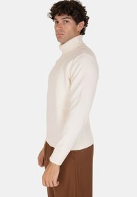 Uomo con i capelli ricci che indossa un maglione a collo alto beige e pantaloni marroni, in piedi di profilo su uno sfondo semplice.