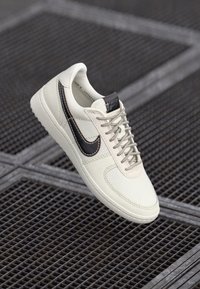 Hvide lædersneakers med mørkebrune swoosh, flade snørebånd og struktureret sål, der har enkle syninger og et minimalistisk design.