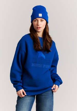 Woman wearing a blue beanie and oversized blue sweatshirt with blue star and text "SNOW MUCH FUN," paired with blue jeans, against plain background.