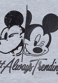 Camiseta gris con dos diseños de Mickey Mouse: uno con un acabado de lentejuelas negras y el otro en un sencillo estampado negro. Texto: "#SiempreEnTendencia."