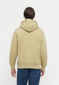 Mann med brunt hår, iført en lys beige hettegenser og blå jeans, som står vendt bort fra kameraet mot en ensfarget hvit bakgrunn.