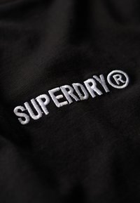 Sort stof med hvid broderet tekst "SUPERDRY" med et registreret varemærkesymbol. Teksturen er glat og let mat.