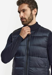 Marinblå quiltad puffer-väst med hög ribbad krage och framdragkedja; i kombination med en mörk långärmad tröja. Slät tygtextur.