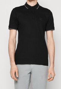Homme portant un polo noir à manches courtes avec un bord gris au col et aux manches, associé à un pantalon gris clair, debout devant un fond uni.