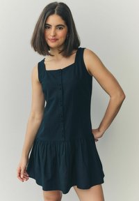 Rochie neagră fără mâneci, cu decolteu pătrat, nasturi frontali și tiv voluminos cu volane. Fabricată dintr-un material ușor, cu o textură netedă.