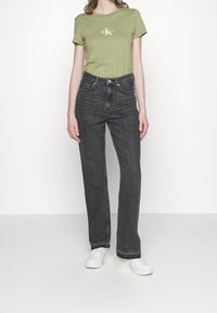 T-shirt en coton vert avec logo, associé à un jean noir droit taille haute et des baskets blanches. Design simple sans motifs visibles.
