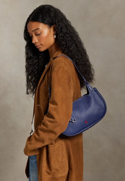 Donna con capelli lunghi e ricci che indossa una giacca marrone e jeans blu, con una borsa a tracolla blu texturizzata con un piccolo logo rosso.