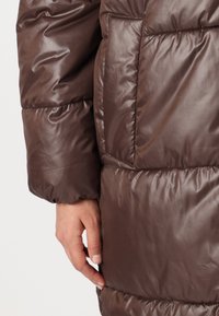 Chaqueta acolchada marrón con un acabado brillante, textura acolchada y un bolsillo lateral. La manga tiene un puño fruncido para un ajuste ceñido.