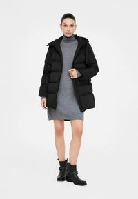 Čierny prešívaný kabát s kapucňou a elastickými manžetami, nosený cez sivé pletené šaty. Osoba má na sebe čierne členkové čižmy s prackovými detailmi.