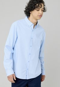 Chemise à manches longues bleu clair, col boutonné, tissu doux, coupe standard, avec des boutons à l'avant et des détails aux poignets, présentant une texture lisse.