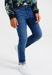 Jeans skinny índigo con cintura media, con un diseño de cinco bolsillos y costuras sutiles. Combinados con zapatillas blancas y dobladillo vuelto.
