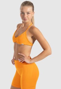 Sujetador deportivo naranja y pantalones cortos de talle alto en una tela suave y elástica. Presenta un centro fruncido y una etiqueta con el logo en los pantalones cortos.