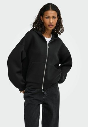 Ung kvinde med mellemtonet hud og krøllet mørkt hår iført en sort hættetrøje med lynlås og mørke jeans, der står mod en ensfarvet lys baggrund.