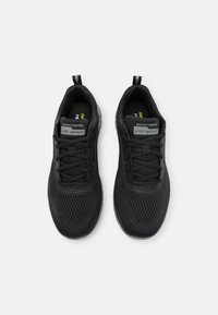 Schwarze Sportschuhe mit einem Mesh-Obermaterial, strukturierten Schnürsenkeln und einer leichten Sohle. Auffälliges Branding auf der Zunge.